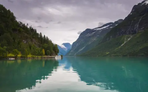 Turkusowa woda odbija góry i lasy, tworząc malowniczy krajobraz norweskich fiordów. Statek cumuje przy brzegu.