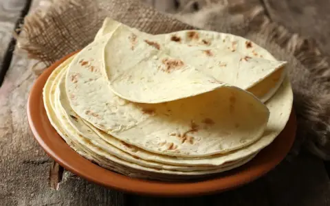 Stos cienkich, jasnych placków keto tortilla na brązowym talerzu, ułożonych na płótnie i drewnianym blacie.