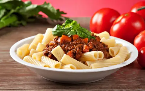 Pyszny makaron włoski rigatoni z sosem bolognese i świeżą bazylią, obok dojrzałe pomidory.