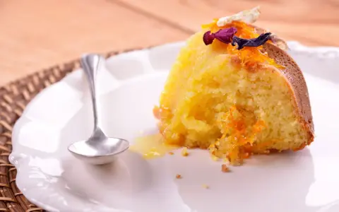 Un trozo de bizcocho de almendra de la abuela, decorado con ralladura de naranja y flores secas, servido en un plato blanco con una cucharilla.