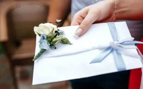 Dłoń trzyma białą kopertę z błękitną wstążką i ozdobnym kwiatkiem. Rozważasz skutki wręczenia pustej koperty lub zbyt małej kwoty? Opinie i etyka są tu kluczowe.