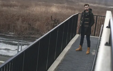 Mężczyzna w budrysówce i traperach stoi na moście z widokiem na zarośla.