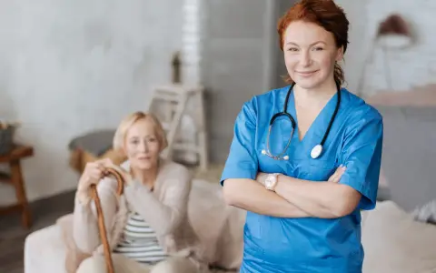 Uśmiechnięta pielęgniarka w niebieskim uniformie, z pewnością ukończyła szybki kurs opiekuna medycznego. W tle starsza pani z laską.