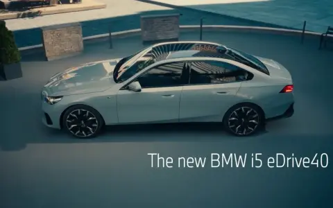 Jasnoszary sedan BMW i5 eDrive40 stoi na zewnątrz, z widokiem na basen i ławkę.
