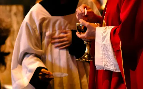 A priest in red vestments holds a chalice and host, while another in white vestments holds a paten. A moment of communion, perhaps a prayer to St. Benedict for healing.