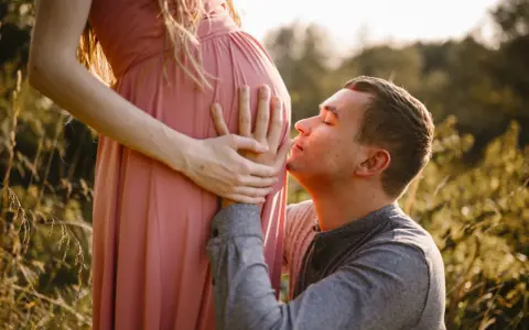 Ciepła sesja ciążowa z mężem. On klęczy i całuje brzuszek ukochanej, otoczeni złotym światłem i naturą.
