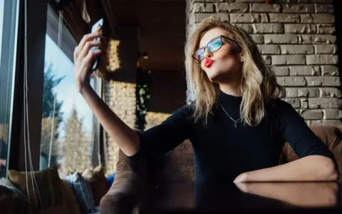 Kobieta w okularach robi sobie selfie, wyciągając usta do pocałunku. W jej spojrzeniu widać pewność siebie, graniczącą z narcyzmem.