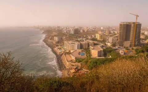 Widok na wybrzeże i rozwijające się miasto w **Afryce, Senegal**. Widać ocean, plażę, nowoczesne budynki i plac budowy z dźwigiem.