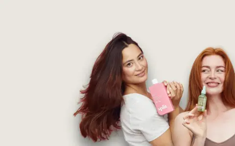 Kobiety prezentują kosmetyki do włosów. Jedna trzyma różową butelkę, druga małą buteleczkę z serum, które zawiera olejek jojoba właściwości odżywcze.