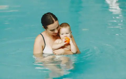 Mama trzyma roczne dziecko na basenie. Maluch bawi się pomarańczową piłeczką.