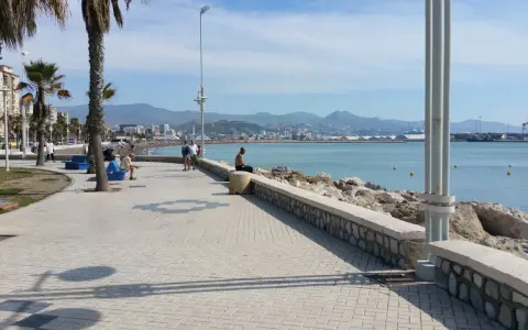 Ein Mann sitzt am Ufer der playa de la misericordia und blickt auf das Meer. Palmen säumen die Promenade, im Hintergrund die Stadt und Berge.