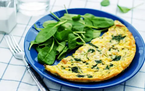 Frittata di spinaci dorata su piatto blu, accompagnata da spinacini freschi. Un pasto leggero e gustoso.