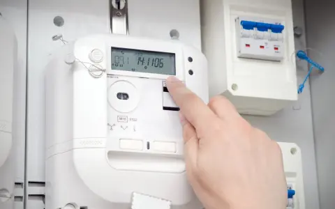 Dłoń naciska przycisk na ekranie inteligentnego licznika energii elektrycznej. Obok widoczny jest wyłącznik.