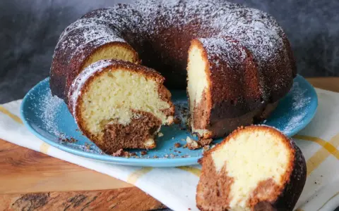 Saftiger weltbester Marmorkuchen, mit Puderzucker bestäubt, auf blauem Teller. Ein Stück ist abgetrennt und zeigt die köstliche Marmorierung.