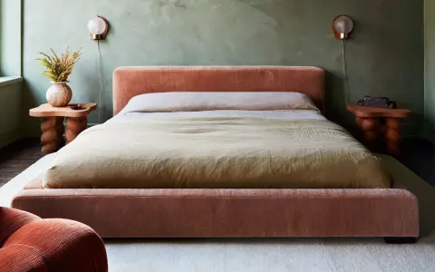 Cama king size tapizada en terciopelo rosa, con mesitas de noche de madera y lámparas de pared.