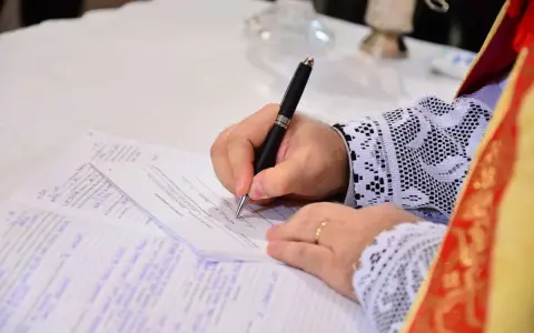 Dłoń w sutannie z koronkowymi mankietami podpisuje akt chrztu do ślubu.