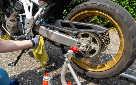 Mano guantata pulisce la catena della moto con uno straccio giallo. Spray per la pulizia catena visibile.