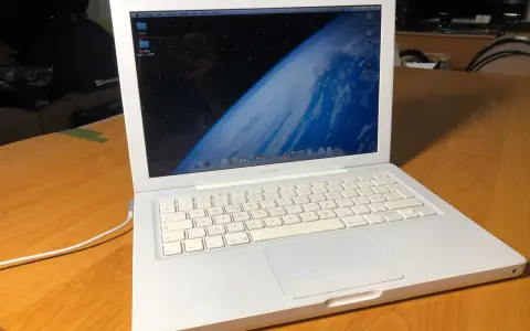Biały, pierwszy MacBook z widokiem Ziemi na ekranie, stoi na drewnianym stole.