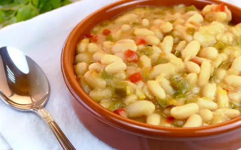 Un cuenco de barro lleno de alubias blancas con verduras, como pimientos rojos y verdes. Una cuchara plateada descansa a la izquierda.