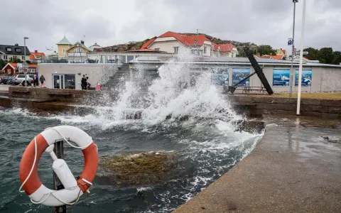Vågor slår mot kajen i en kuststad, med hus i bakgrunden. En livboj syns i förgrunden. Det blåser med sekundmeter storm.