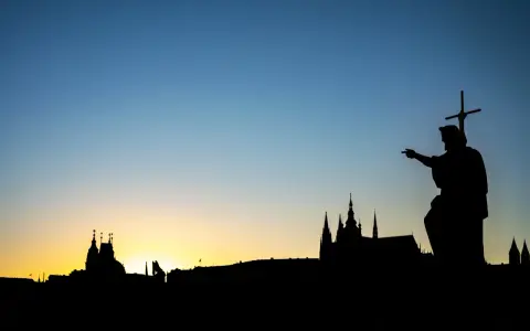 Silhouette d'une statue de saint pointant vers un paysage urbain au coucher du soleil. Le ciel bleu dégradé est le centre d'intérêt de l'image.