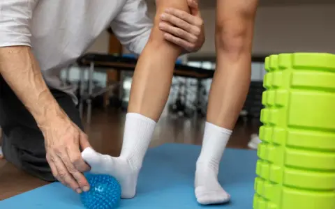 Fizjoterapeuta pomaga w rehabilitacji stopy, masując ją kolczastą piłeczką. Obok zielony wałek do ćwiczeń.