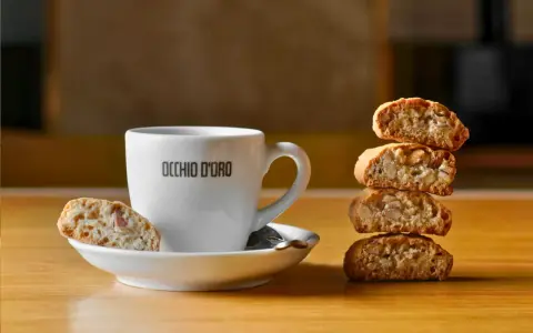 Ein weißer Kaffeebecher mit "OCCHIO D'ORO" Schriftzug steht neben einem Stapel Cantuccini. Ein Cantuccino liegt auf der Untertasse. Dieses Bild inspiriert zum besten Cantuccini Rezept.