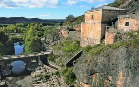 El portón de la oliva, un antiguo edificio de piedra junto a un río y un puente de arco, se alza sobre un paisaje rocoso y verde.