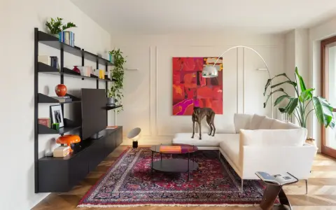 Un galgo se posa sobre un sofá blanco, rodeado de arte abstracto y un tapete persa. El **ruido visual** de los objetos decorativos y la pintura vibrante crea un ambiente ecléctico.