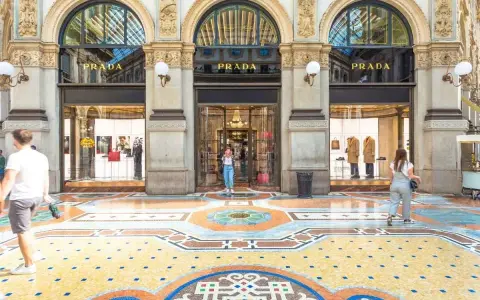 Ingresso del negozio **Prada** con vetrine eleganti e un mosaico colorato sul pavimento. Persone passeggiano davanti alla boutique.