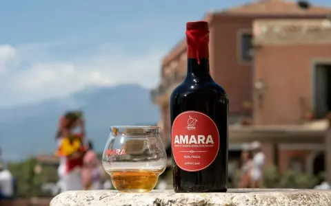 Bottiglia e bicchiere di Amara, un amaro che celebra come l'arancio amaro trama i sapori siciliani. Sfondo sfocato di un borgo.