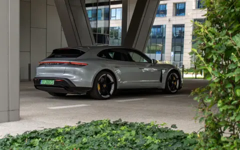 Szare Porsche Taycan Turbo S stoi pod zadaszeniem, z widokiem na nowoczesny budynek i zieleń.