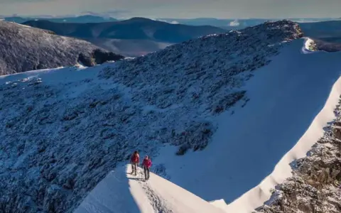 Två vandrare på en snöig bergskam med utsikt över fjällandskap. Detta är Sveriges högsta berg.