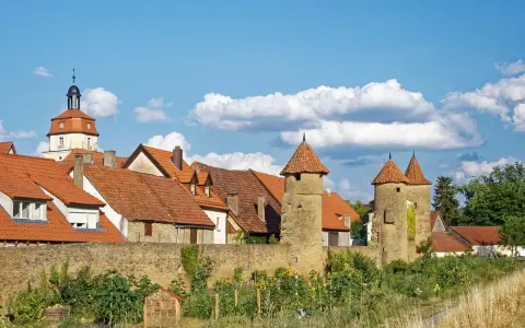 Eine malerische **Stadt im Mittelalter** mit roten Dächern, steinernen Türmen und einer alten Stadtmauer unter blauem Himmel.
