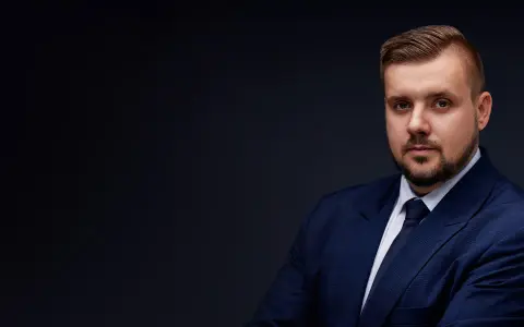 Mężczyzna w granatowym garniturze i krawacie, z zarostem, na ciemnym tle.