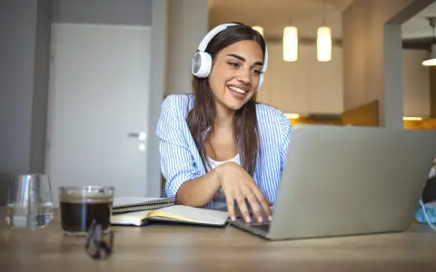 Uśmiechnięta studentka w słuchawkach pracuje przy laptopie. Czy muzyka pomaga w nauce? Ona chyba tak, bo wygląda na zrelaksowaną i skupioną.