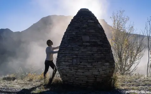 Une femme interagit avec une structure ovoïde en pierres, une œuvre de land art, dans un paysage brumeux et montagneux au lever du soleil.