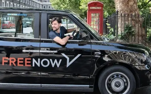 Ein Mann fährt ein schwarzes freenow taxi. Im Hintergrund steht eine rote Telefonzelle.
