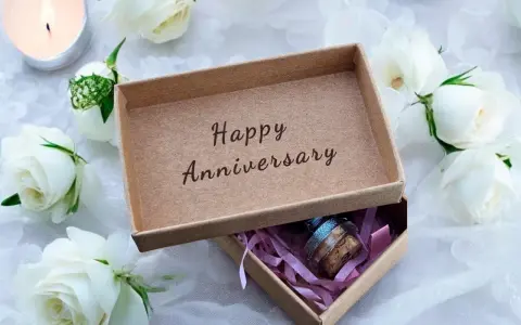 Otwarta szkatułka z napisem "Happy Anniversary" i małą buteleczką w środku, otoczona białymi różami i płonącą świecą. Idealny prezent na 21 rocznicę ślubu.