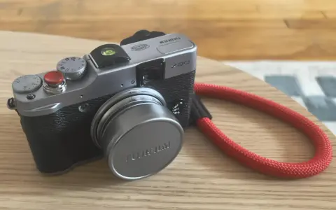 Srebrny aparat Fujifilm X20 z czerwoną smyczą leży na drewnianym blacie.