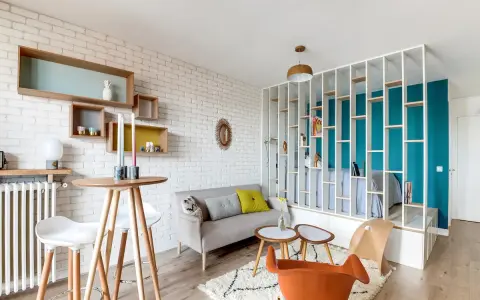 Idée aménagement studio 25m2 : un espace salon cosy avec canapé, table haute et étagères murales, séparé d'une chambre par une bibliothèque ajourée.