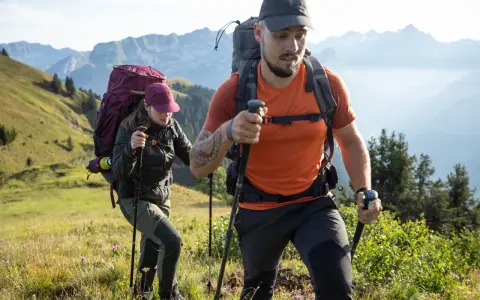 Dos excursionistas con mochilas y bastones caminan por una ladera montañosa.