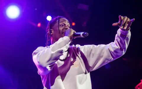 Travis Scott na scenie, w białej bluzie, z mikrofonem w dłoni. Jego energia inspiruje wielu **współautorów piosenek**.