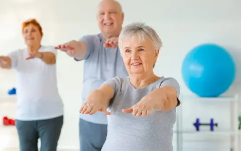 Trois seniors souriants pratiquent la gym douce, bras tendus, dans une salle lumineuse avec un ballon bleu en arrière-plan.