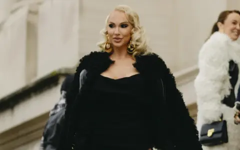 Eine Frau im Stil einer "mob wife" mit blonden Locken, großen goldenen Ohrringen und einem schwarzen Pelzmantel.