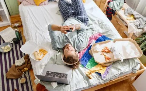 Mann liegt im Chaos auf dem Bett, umgeben von Essensresten und Kleidung. Ein typischer Tag des "bed rotting".