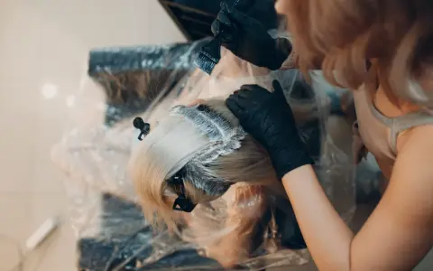 Fryzjerka w czarnych rękawiczkach nakłada farbę na blond włosy. Kluczowe jest, ile trzymać farbę na włosach, by uzyskać idealny efekt.