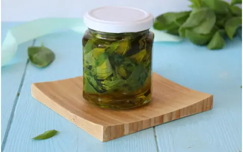 Un barattolo di olio al basilico fatto in casa, con foglie fresche immerse nell'olio dorato, su un tagliere di legno.