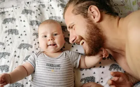 Ein lächelnder Vater und sein Baby liegen auf einer Decke mit Bärenmuster. Die Frage, ob Autismus vererbbar ist, beschäftigt viele, und die Bindung zwischen Vater und Kind ist ein wichtiger Aspekt.