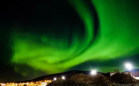 Spektakularna zorza polarna rozświetla nocne niebo nad ośnieżonymi wzgórzami i światłami miasteczka. To idealny moment, kiedy występuje zorza polarna.